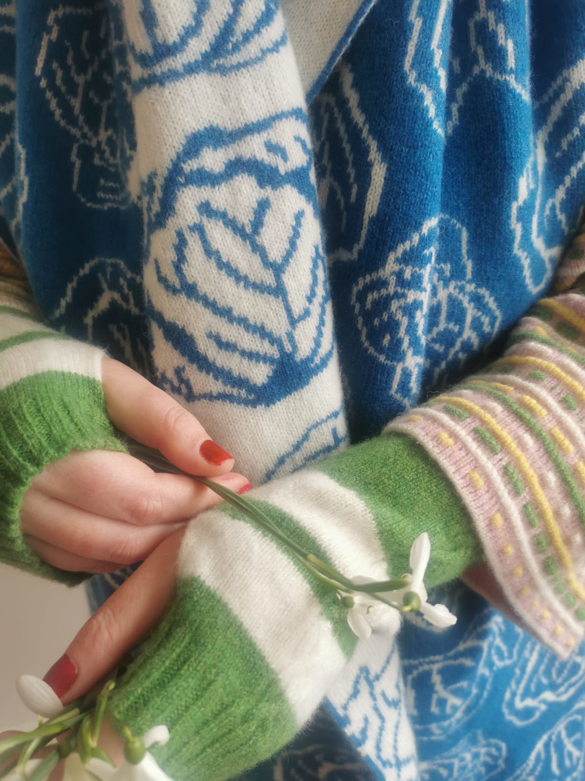 Muddle Bus 03 Pied Piper Wrist Warmers. Hands in front of blue and white knitted scarf with a pattern of cabbages (Muddle Bus 05). Hands wearing Green and white striped knitted fingerless gloves holding snowdrops.