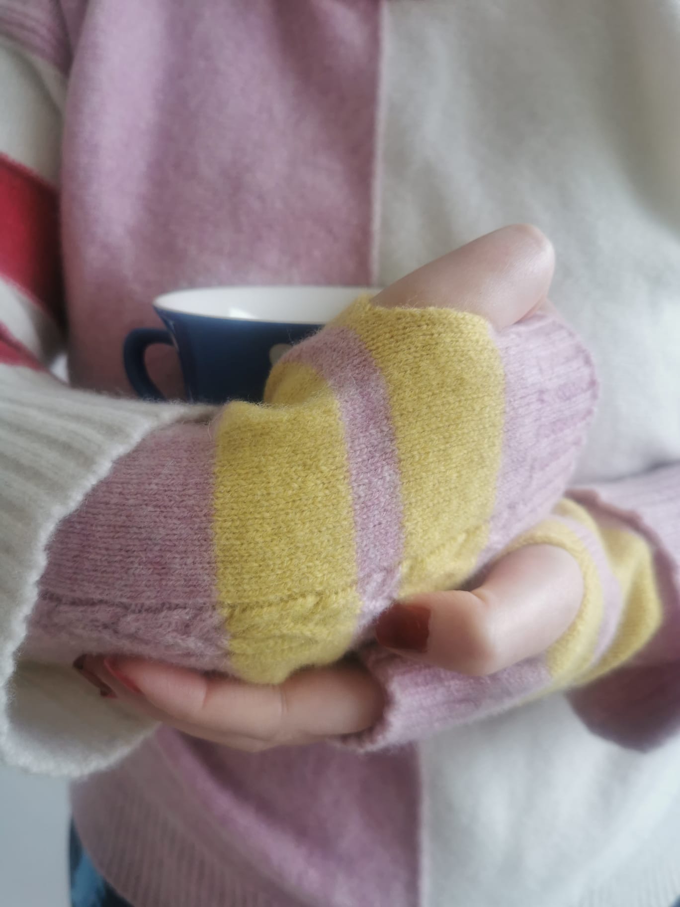 Muddle Bus 03- Pied Piper Wrist Warmers. Hands folded around a blue spot cup wearing knitted pink and yellow stripe fingerless gloves. Muddle Bus 02 Pied Piper Jumper in Pink/Pink/White as background,