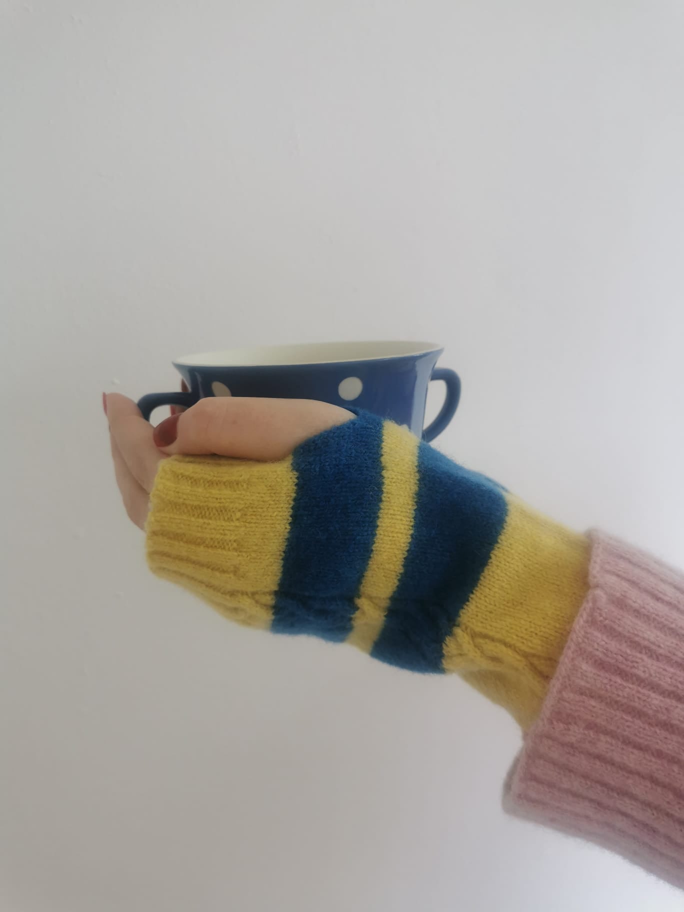 Muddle Bus 03 Pied Piper Fingerless Gloves. Hands wearing yellow and blue striped knitted fingerless gloves holding blue cup with white spots in front of white wall.